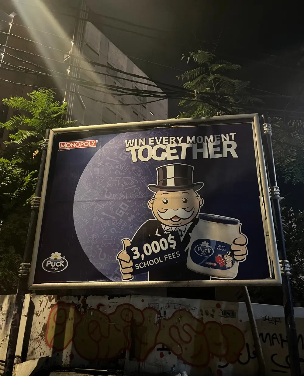 Monopoly co-branded bir yoğurt reklamı reklam panosunda görünüyor. Akşam vaktinin karanlığı ve reklamın siyah arka planı birbirine geçişli.